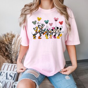 May include: Pink t-shirt with a graphic print of Mickey Mouse, Minnie Mouse, and Goofy dressed in skeleton costumes holding colorful balloons. The characters are smiling and appear to be dancing.