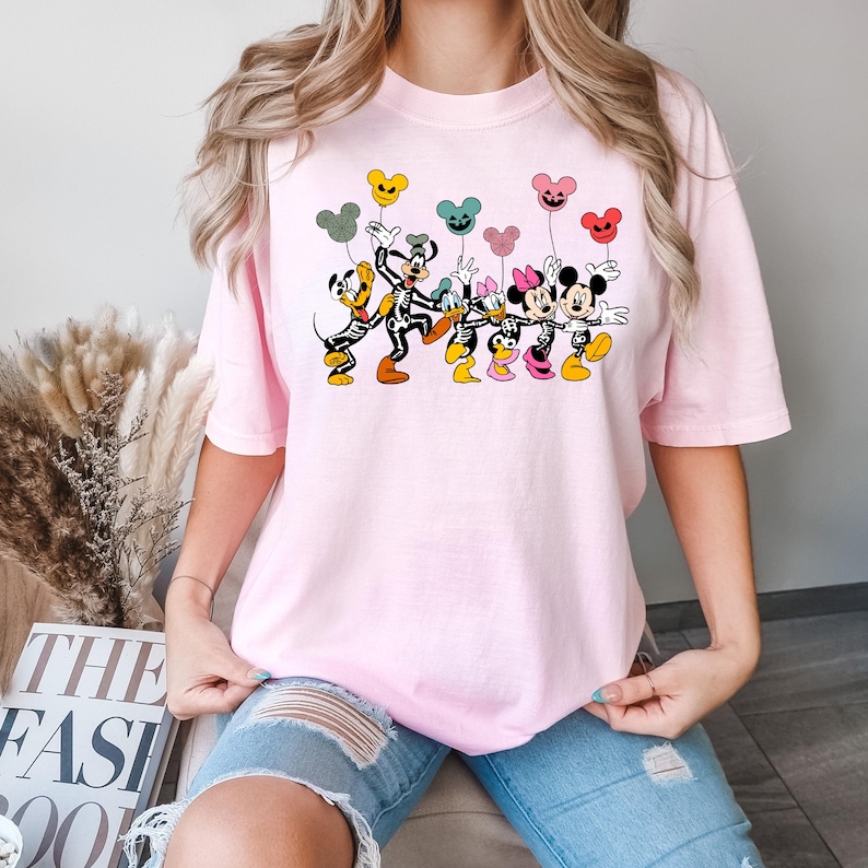 May include: Pink t-shirt with a graphic print of Mickey Mouse, Minnie Mouse, and Goofy dressed in skeleton costumes holding colorful balloons. The characters are smiling and appear to be dancing.