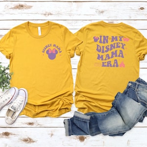 May include: Mustard yellow t-shirts with the text "Disney Mama" and a Minnie Mouse graphic on the front. The back of the shirt says "In My Disney Mama Era" with a castle graphic. Blue jeans and white trainers are also in the image.