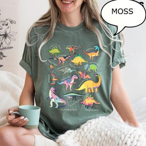 May include: A moss green t-shirt featuring a colorful dinosaur graphic. The graphic includes various dinosaur illustrations with their names. The shirt is worn by a person holding a blue mug.