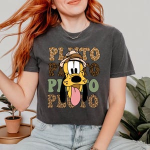 May include: A dark grey cropped t-shirt featuring a cartoon image of Pluto wearing a safari hat. The shirt has a repeating pattern of leopard print, giraffe print, and green geometric shapes. The text "PLUTO" is repeated in a stylised font.