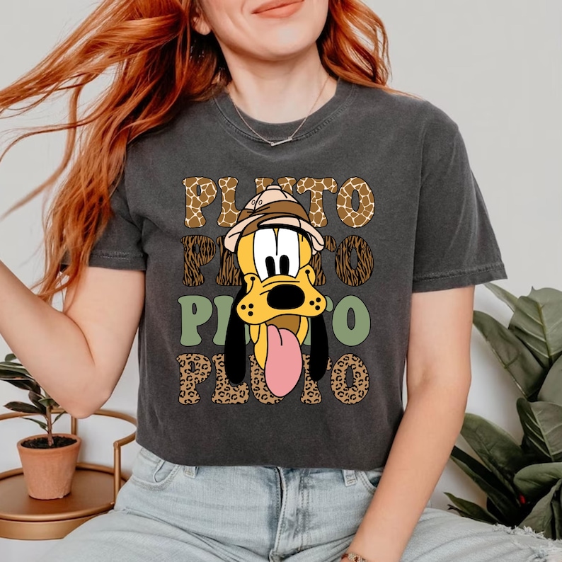 May include: A dark grey cropped t-shirt featuring a cartoon image of Pluto wearing a safari hat. The shirt has a repeating pattern of leopard print, giraffe print, and green geometric shapes. The text "PLUTO" is repeated in a stylised font.