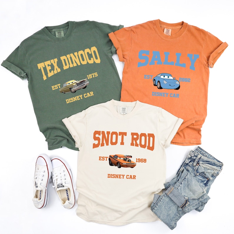 Puede incluir: Tres camisetas con dise&ntilde;os de personajes de la pel&iacute;cula Cars. La camiseta verde dice "Tex Dinoco Est. 1975 Disney Car" con una imagen de un coche amarillo. La camiseta naranja dice "Sally Est. 2002 Disney Car" con una imagen de un coche azul. La camiseta beige dice "Snot Rod Est. 1968 Disney Car" con una imagen de un coche naranja.