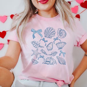 May include: A pink t-shirt with a blue and white seashell design. The design features various seashells, starfish, and bows.