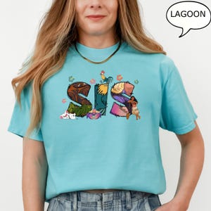 May include: A turquoise blue t-shirt featuring a graphic of the word "SIS" made up of images from the Disney film Moana. The letters are decorated with flowers, a rooster, a crab, and a character from the film.