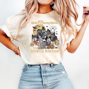 Puede incluir: Una camiseta blanca con un gr&aacute;fico de estilo vintage de Mickey Mouse y sus amigos frente a la Mansi&oacute;n Encantada en Walt Disney World. El texto "Walt Disney World" y "The Haunted Mansion" est&aacute; impreso en dorado en la camiseta.