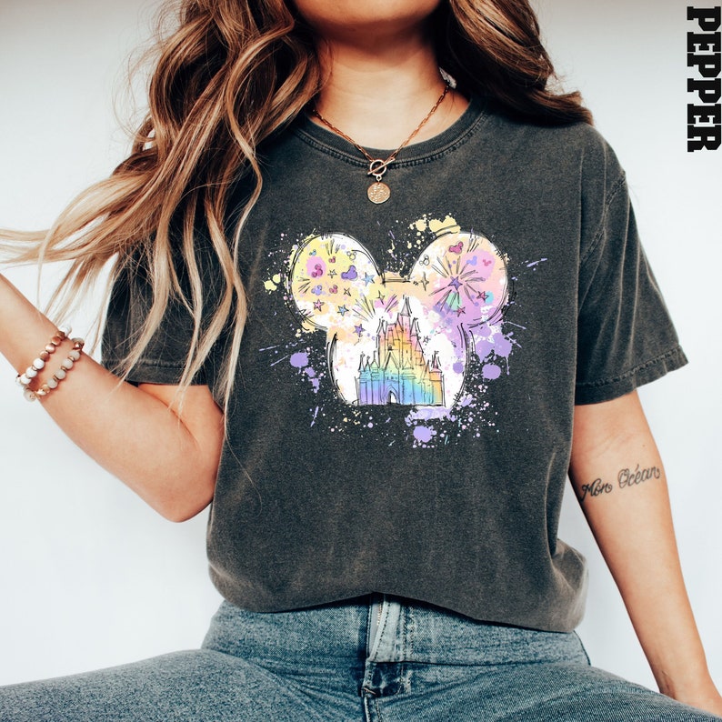 Puede incluir: Una camiseta gris oscuro con un gr&aacute;fico colorido de un castillo en forma de oreja de Mickey Mouse. El gr&aacute;fico tiene un efecto acuarela e incluye estrellas, corazones y fuegos artificiales.