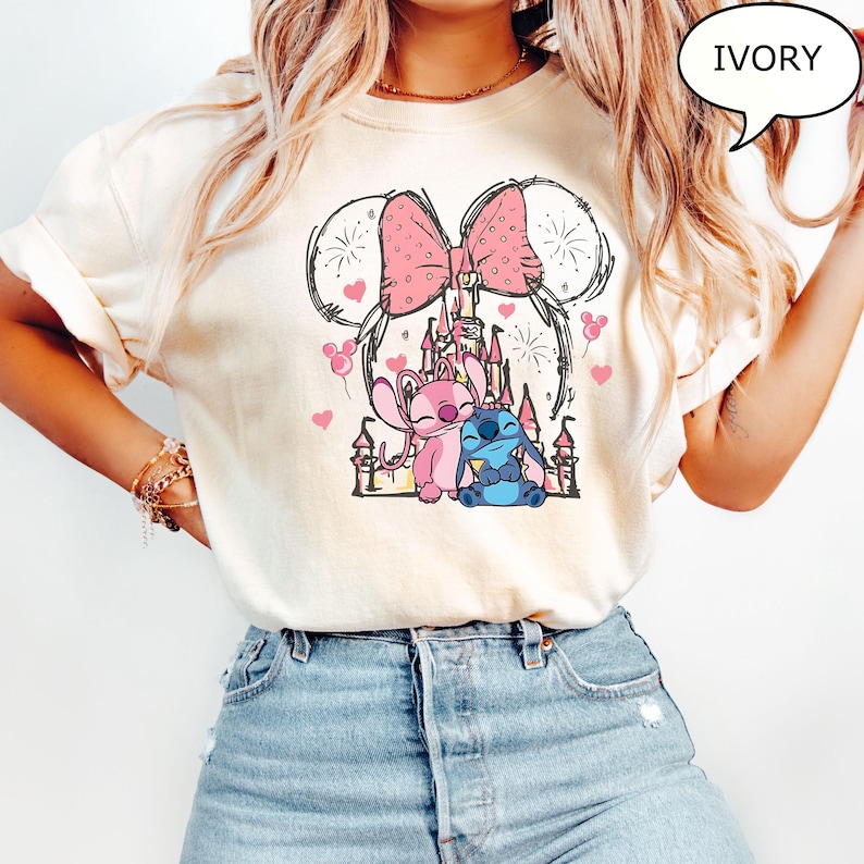 Puede incluir: Camiseta de color marfil con un gr&aacute;fico de dibujos animados de un lazo rosa con lunares, orejas de Mickey Mouse y un castillo. El gr&aacute;fico incluye corazones rosas, fuegos artificiales y dos personajes de dibujos animados. La palabra "IVORY" est&aacute; en una burbuja.