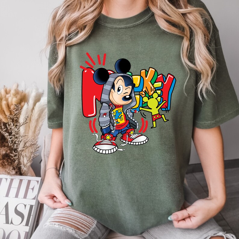 Pode incluir: Uma camiseta verde com uma imagem de desenho animado do Mickey Mouse vestindo um moletom com capuz e t&ecirc;nis vermelhos. O texto "Mickey" est&aacute; escrito em letras coloridas ao redor do Mickey Mouse.