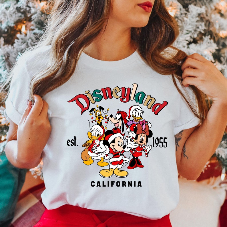 May include: White t-shirt with a colorful Disneyland logo featuring Mickey Mouse, Minnie Mouse, Donald Duck, Goofy, and Pluto wearing Santa hats. The text reads "Disneyland est. 1955 CALIFORNIA".
