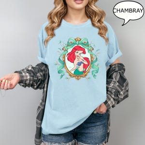 May include: Light blue t-shirt featuring a cartoon image of Ariel from The Little Mermaid, framed by a gold oval and teal ribbon. The word "CHAMBRAY" is in a speech bubble. The shirt is a light blue color.