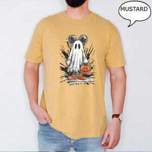May include: Mustard-colored t-shirt featuring a Halloween-themed graphic. The graphic depicts a ghost with Mickey Mouse ears holding a jack-o'-lantern bucket. The word "MUSTARD" is in a speech bubble. The ghost is wearing orange shoes.