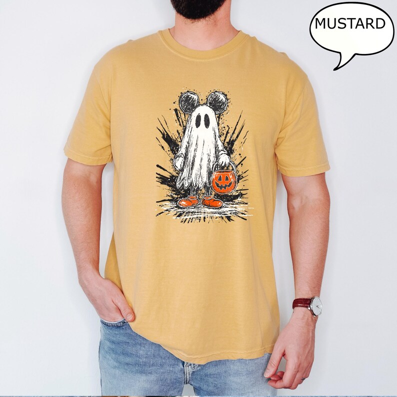 May include: Mustard-colored t-shirt featuring a Halloween-themed graphic. The graphic depicts a ghost with Mickey Mouse ears holding a jack-o'-lantern bucket. The word "MUSTARD" is in a speech bubble. The ghost is wearing orange shoes.