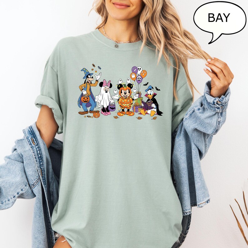 May include: A light green t-shirt featuring a Halloween-themed graphic of Disney characters, including Mickey Mouse, Minnie Mouse, Donald Duck, and Goofy, dressed in costumes. The word "BAY" is in a speech bubble.