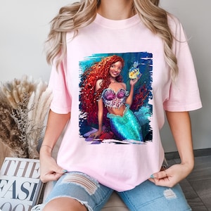 May include: A pink t-shirt featuring a graphic print of a mermaid with long red hair holding a yellow fish. The mermaid is wearing a seashell bra and a green tail.