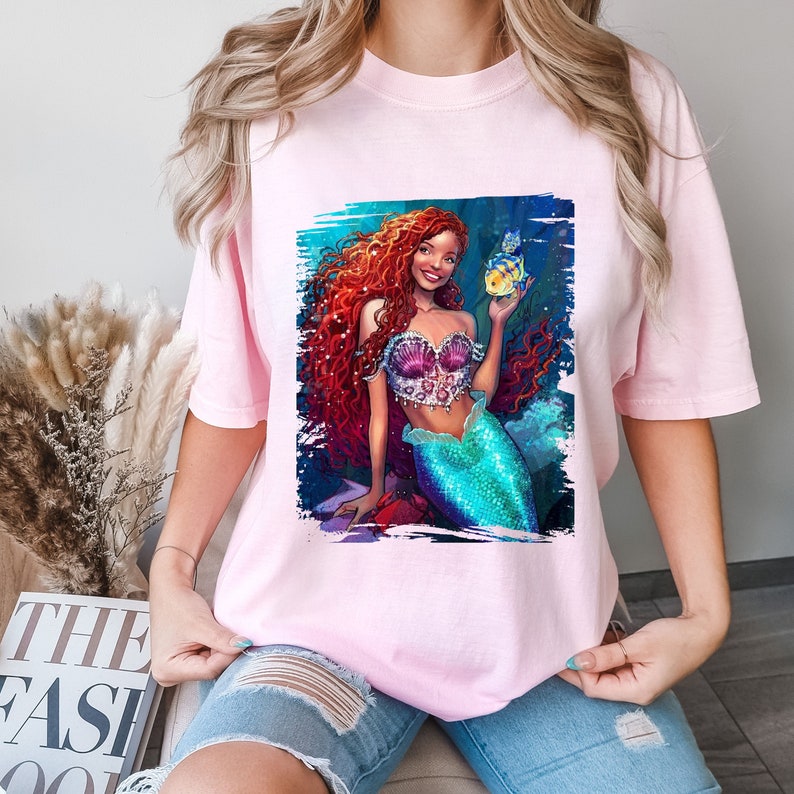 May include: A pink t-shirt featuring a graphic print of a mermaid with long red hair holding a yellow fish. The mermaid is wearing a seashell bra and a green tail.