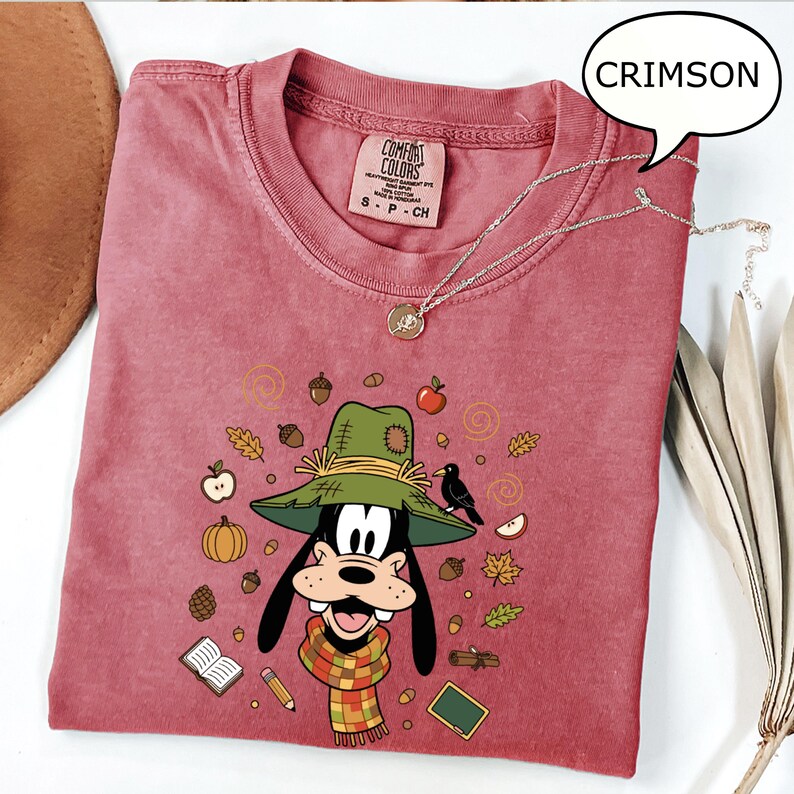 May include: Crimson-colored t-shirt featuring a cartoon character wearing a green hat and a plaid scarf, surrounded by fall-themed illustrations. The shirt has a crew neck and a small tag. A silver necklace with a pendant is draped across the shirt.