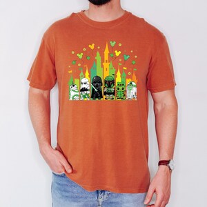 Puede incluir: Una camiseta naranja con un gr&aacute;fico colorido de personajes de Star Wars frente a un castillo. Los personajes incluyen un Grogu verde, un Stormtrooper, Darth Vader, Boba Fett y R2-D2. Tr&eacute;boles verdes y orejas de Mickey Mouse tambi&eacute;n est&aacute;n en el dise&ntilde;o.