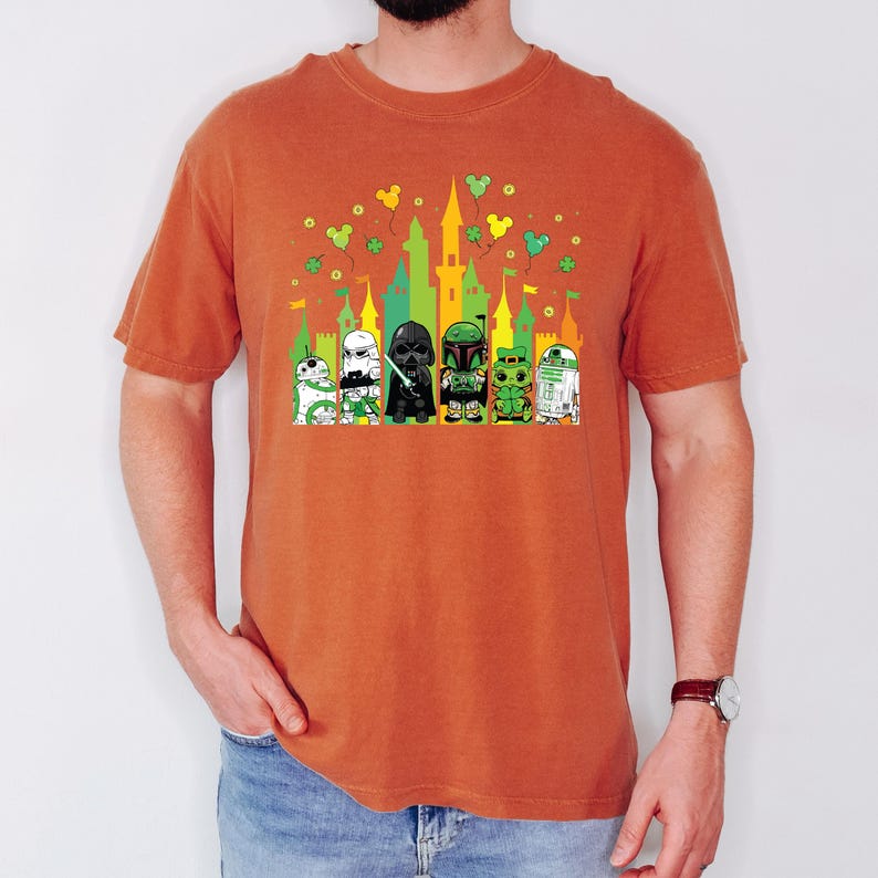 Puede incluir: Una camiseta naranja con un gr&aacute;fico colorido de personajes de Star Wars frente a un castillo. Los personajes incluyen un Grogu verde, un Stormtrooper, Darth Vader, Boba Fett y R2-D2. Tr&eacute;boles verdes y orejas de Mickey Mouse tambi&eacute;n est&aacute;n en el dise&ntilde;o.