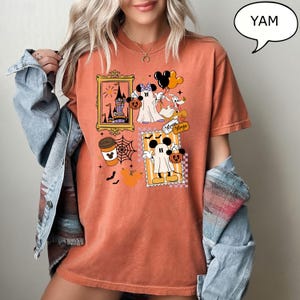 Może przedstawiać: Pomarańczowa koszulka z grafiką o tematyce Halloween. Projekt zawiera obrazy Myszki Miki i Myszki Minnie przebranych za duchy, filiżankę kawy, pajęczynę i zamek. Koszulka zawiera r&oacute;wnież napis "Minnie Magic".