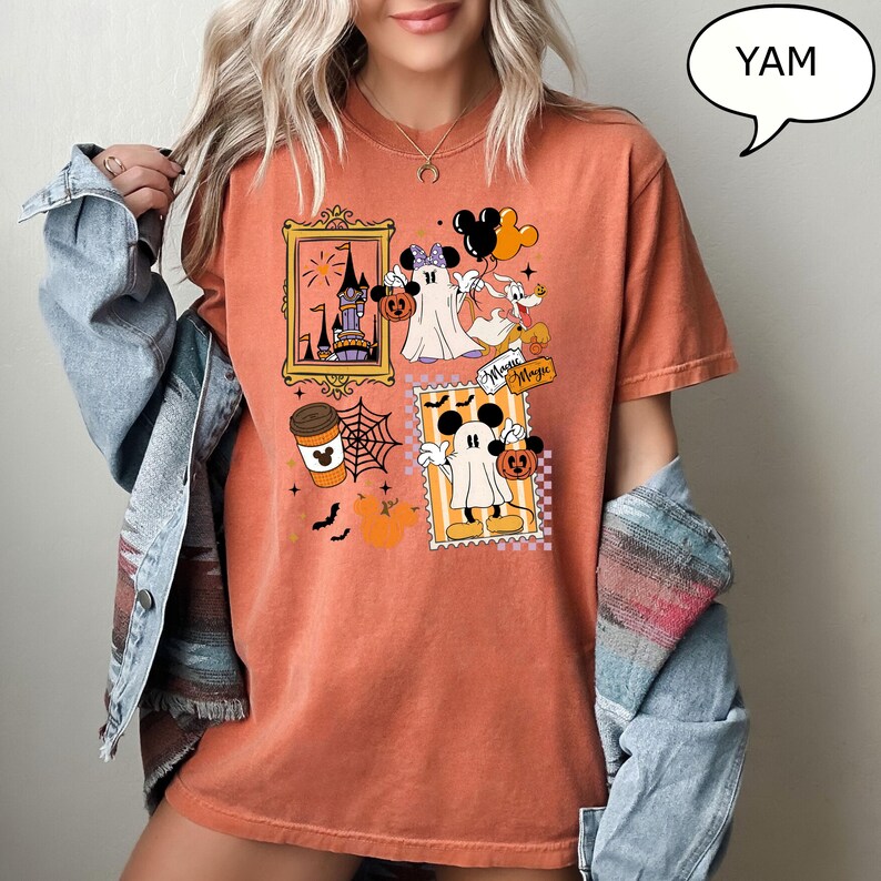 Może przedstawiać: Pomarańczowa koszulka z grafiką o tematyce Halloween. Projekt zawiera obrazy Myszki Miki i Myszki Minnie przebranych za duchy, filiżankę kawy, pajęczynę i zamek. Koszulka zawiera r&oacute;wnież napis "Minnie Magic".