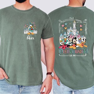 May include: Green t-shirt with a vintage-style graphic of Mickey Mouse, Minnie Mouse, Donald Duck, Goofy, and Pluto in front of Cinderella's Castle. The text "Disneyworld Est. 1965" is below the characters. The front of the shirt has a smaller graphic of Donald Duck in front of Cinderella's Castle with the name "Alex" below.