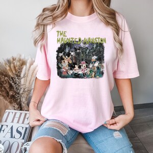 May include: Pink t-shirt with a graphic print of Mickey Mouse and friends in front of a haunted house. The text "The Haunted Mansion" is printed in green on the shirt.
