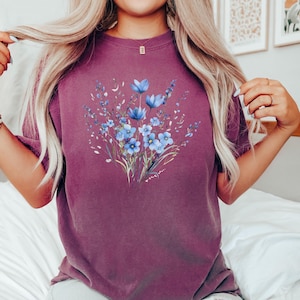 Może przedstawiać: Fioletowy t-shirt z niebieskim wzorem kwiatowym. Kwiaty są w stylu akwarelowym i mają jasnoniebieski kolor.
