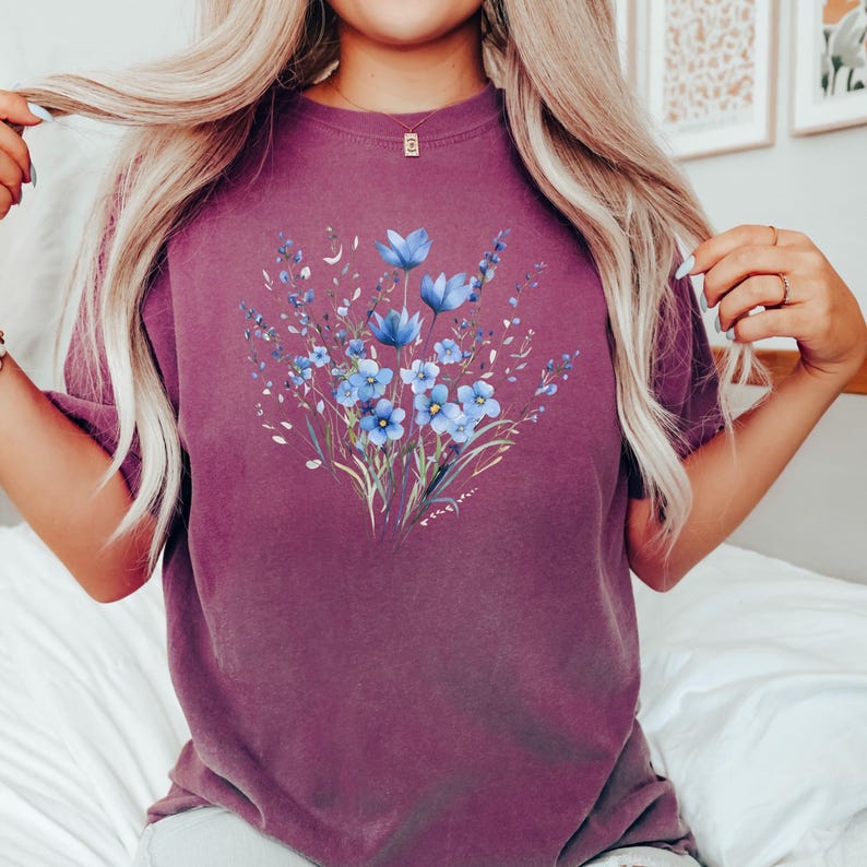 Może przedstawiać: Fioletowy t-shirt z niebieskim wzorem kwiatowym. Kwiaty są w stylu akwarelowym i mają jasnoniebieski kolor.