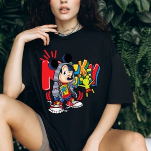 Pode incluir: Camiseta preta com um gr&aacute;fico colorido do Mickey Mouse vestindo um moletom com capuz e jeans. O gr&aacute;fico inclui a palavra "Mickey" em uma fonte colorida de estilo graffiti.