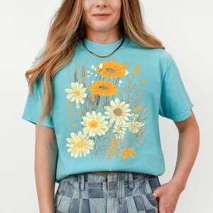 May include: A light blue t-shirt featuring a floral design with yellow and white daisies and orange sunflowers. The graphic includes various other floral elements in shades of beige and brown. The shirt has short sleeves and a crew neck.