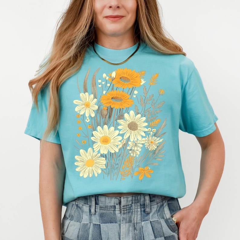 May include: A light blue t-shirt featuring a floral design with yellow and white daisies and orange sunflowers. The graphic includes various other floral elements in shades of beige and brown. The shirt has short sleeves and a crew neck.