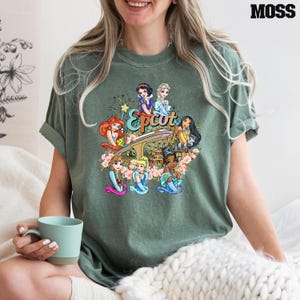 May include: A green t-shirt featuring a graphic of Disney princesses in front of the Epcot logo. The princesses include Snow White, Elsa, Ariel, Pocahontas, and Mulan.