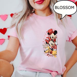 Puede incluir: Camiseta rosa claro con Mickey Mouse, Minnie Mouse y Pluto con atuendos de San Valent&iacute;n. Mickey lleva un sombrero de copa, Minnie un lazo y Pluto un collar de reno. La palabra "BLOSSOM" est&aacute; en una burbuja.