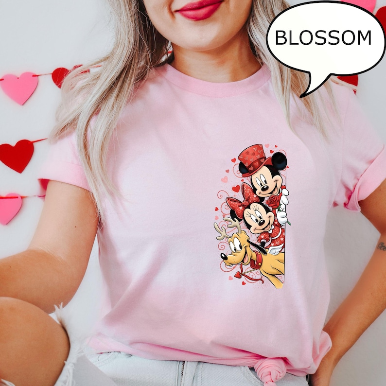Puede incluir: Camiseta rosa claro con Mickey Mouse, Minnie Mouse y Pluto con atuendos de San Valent&iacute;n. Mickey lleva un sombrero de copa, Minnie un lazo y Pluto un collar de reno. La palabra "BLOSSOM" est&aacute; en una burbuja.