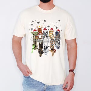 Puede incluir: Una camiseta blanca con un gr&aacute;fico de Star Wars con tem&aacute;tica navide&ntilde;a. El gr&aacute;fico presenta personajes como Yoda, Darth Vader, R2-D2 y Chewbacca con gorros de Pap&aacute; Noel y reflejados en un charco de agua.