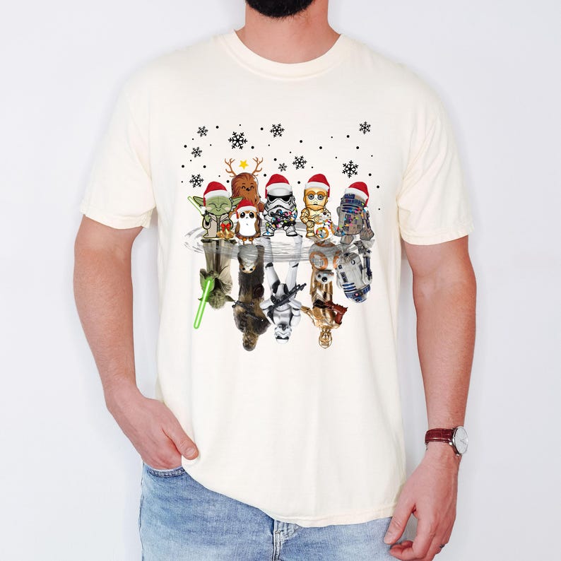 Puede incluir: Una camiseta blanca con un gr&aacute;fico de Star Wars con tem&aacute;tica navide&ntilde;a. El gr&aacute;fico presenta personajes como Yoda, Darth Vader, R2-D2 y Chewbacca con gorros de Pap&aacute; Noel y reflejados en un charco de agua.