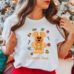 May include: White t-shirt with a gingerbread mouse character wearing a bow. The character is surrounded by Christmas-themed decorations like candy canes, bells, and gift boxes. The text "Custom Name" is printed below the character.