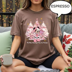 May include: Espresso-colored t-shirt featuring a Minnie Mouse graphic in a Christmas theme. Minnie is dressed in pink, holding a gift, with pink Christmas trees in the background. The text "The Most Wonderful Time Of The Year" is printed below the graphic.