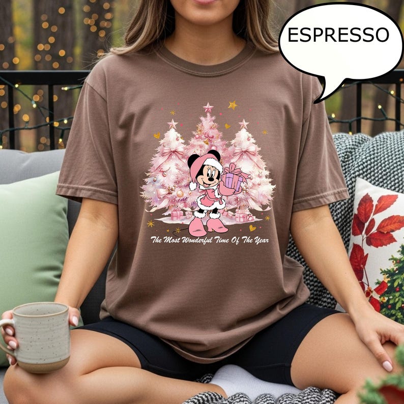 May include: Espresso-colored t-shirt featuring a Minnie Mouse graphic in a Christmas theme. Minnie is dressed in pink, holding a gift, with pink Christmas trees in the background. The text "The Most Wonderful Time Of The Year" is printed below the graphic.