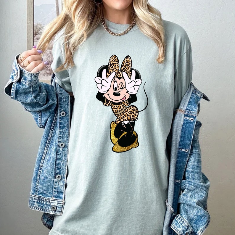 K&ouml;nnte beinhalten: Hellgr&uuml;nes T-Shirt mit einer Cartoon-Figur von Minnie Mouse in einem Leopardenmuster-Outfit. Minnie macht mit beiden H&auml;nden ein Friedenszeichen. Ihre Schuhe sind goldfarben glitzernd. Das Shirt hat eine lockere Passform.