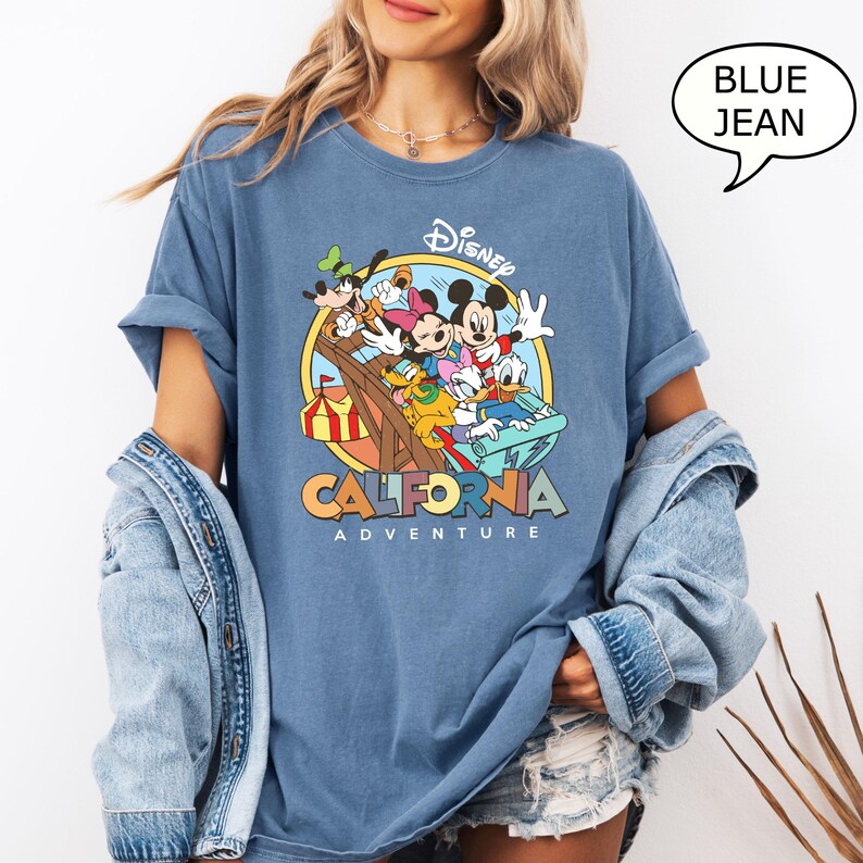 Puede incluir: Una camiseta azul con un gr&aacute;fico de Mickey Mouse y sus amigos montando una monta&ntilde;a rusa en Disney California Adventure. El texto "Disney California Adventure" est&aacute; impreso debajo del gr&aacute;fico.