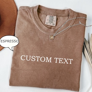 May include: A brown t-shirt with the text "CUSTOM TEXT" printed in white. The shirt has a distressed look and is made of a soft, comfortable fabric. The shirt is shown folded on a white surface.