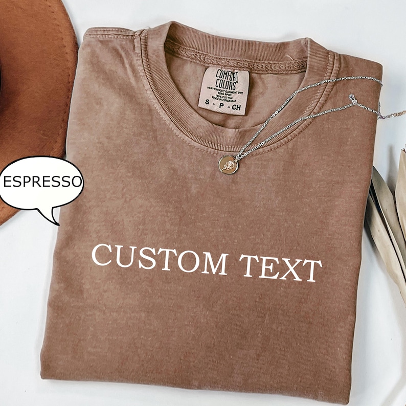 May include: A brown t-shirt with the text "CUSTOM TEXT" printed in white. The shirt has a distressed look and is made of a soft, comfortable fabric. The shirt is shown folded on a white surface.