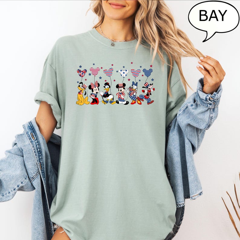 Puede incluir: Camiseta verde salvia con un gr&aacute;fico de personajes de Disney sosteniendo globos en forma de coraz&oacute;n rojos, blancos y azules. Los personajes incluyen a Pluto, Minnie Mouse, Donald Duck, Mickey Mouse y Goofy. La palabra "BAY" est&aacute; en una burbuja.