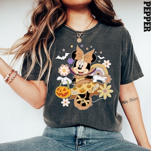 Puede incluir: Camiseta gris oscuro con un dise&ntilde;o de Minnie Mouse de Halloween. Minnie est&aacute; disfrazada, sosteniendo una linterna de calabaza, con un arco&iacute;ris y flores. La camiseta tiene un aspecto vintage y desgastado.