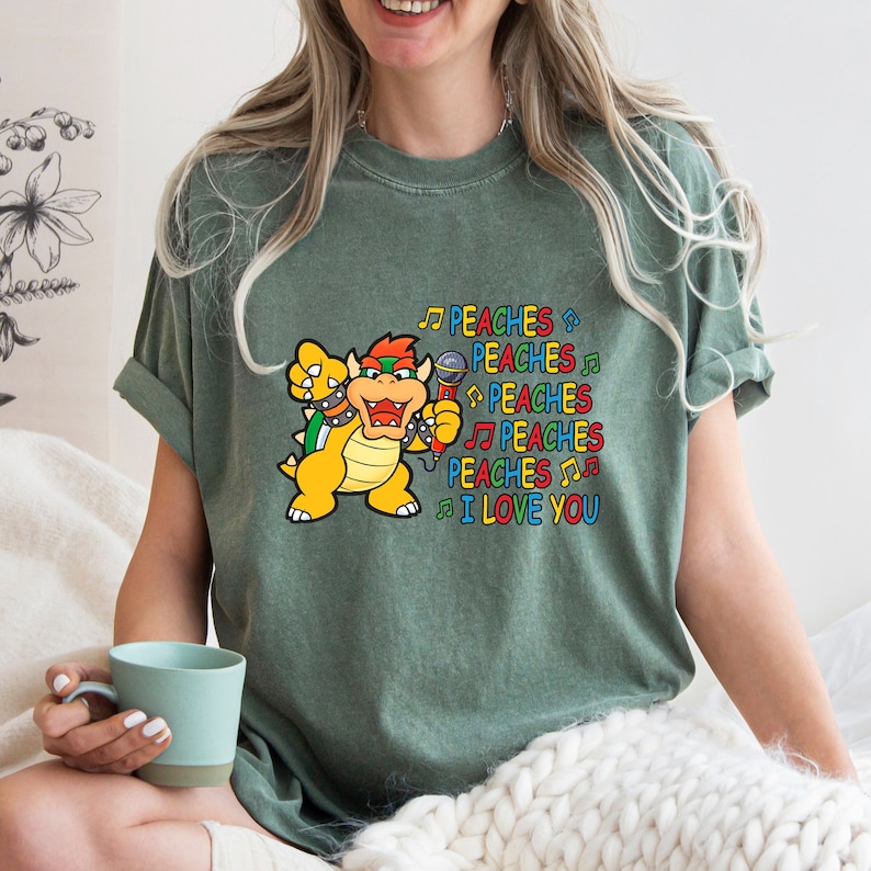 May include: Green t-shirt featuring a cartoon Bowser character holding a microphone. The text "PEACHES I LOVE YOU" is printed in colorful letters and musical notes. The shirt is a comfortable, casual style.