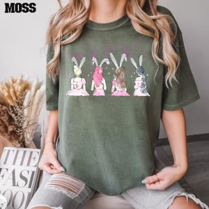 Puede incluir: Camiseta verde con un gr&aacute;fico de cuatro chicas con orejas de conejo. Las chicas llevan vestidos rosas y tienen el pelo de diferentes colores. El gr&aacute;fico es de estilo de dibujos animados.