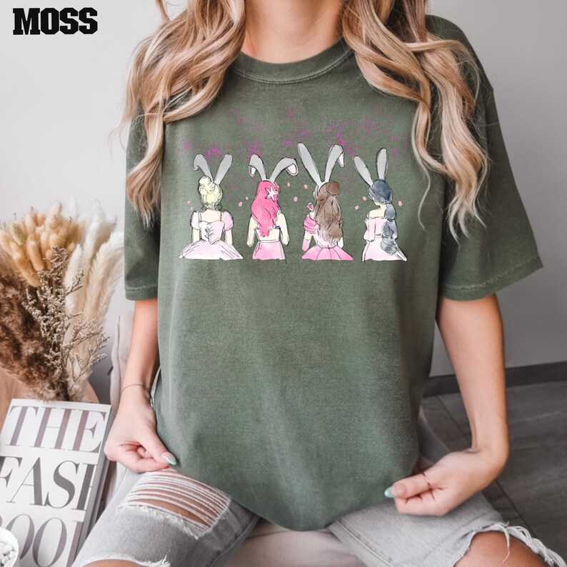 Puede incluir: Camiseta verde con un gr&aacute;fico de cuatro chicas con orejas de conejo. Las chicas llevan vestidos rosas y tienen el pelo de diferentes colores. El gr&aacute;fico es de estilo de dibujos animados.