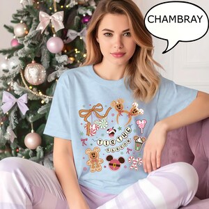 May include: Light blue t-shirt with a festive graphic featuring gingerbread men, candy canes, and the phrase "Tis the Season." The shirt is paired with striped pajama pants, creating a cozy holiday ensemble. The word "CHAMBRAY" is in a speech bubble.